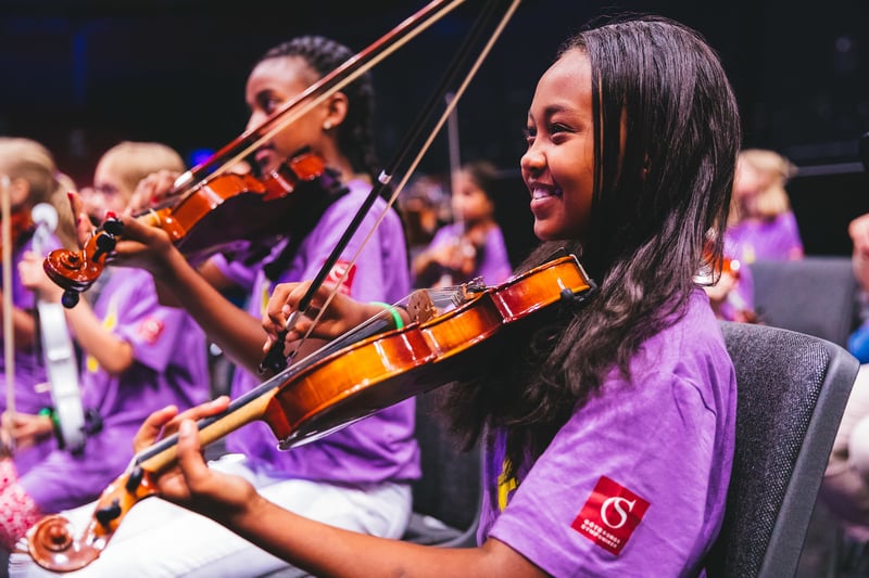 Barn i lila t-shirtar som spelar i stråkorkester. I förgrunden en leende flicka som håller i sin violin.
